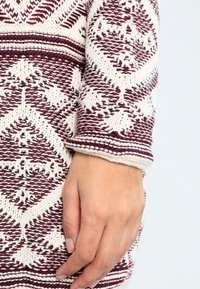 Indumento lavorato a maglia con colori marrone e crema, intricati motivi geometrici, tessuto testurizzato e primo piano della manica con polsino.