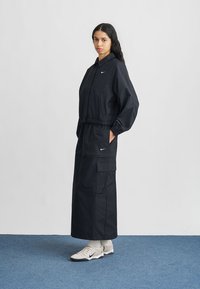 Μαύρο δύο τεμαχίων σύνολο που περιλαμβάνει ζακέτα με φερμουάρ και ελαστικές μανσέτες και μακριά φούστα με τσέπες cargo, σε συνδυασμό με λευκά αθλητικά παπούτσια.