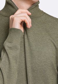 Pull en zip couleur vert olive avec un col montant, des poignets côtelés et un tricot texturé. La fermeture éclair possède un tirette en métal.
