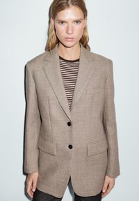 Blazer in lana beige con spalle strutturate, rever a punta e due tasche frontali. Indossato sopra una camicia a righe scure, con bottoni neri.