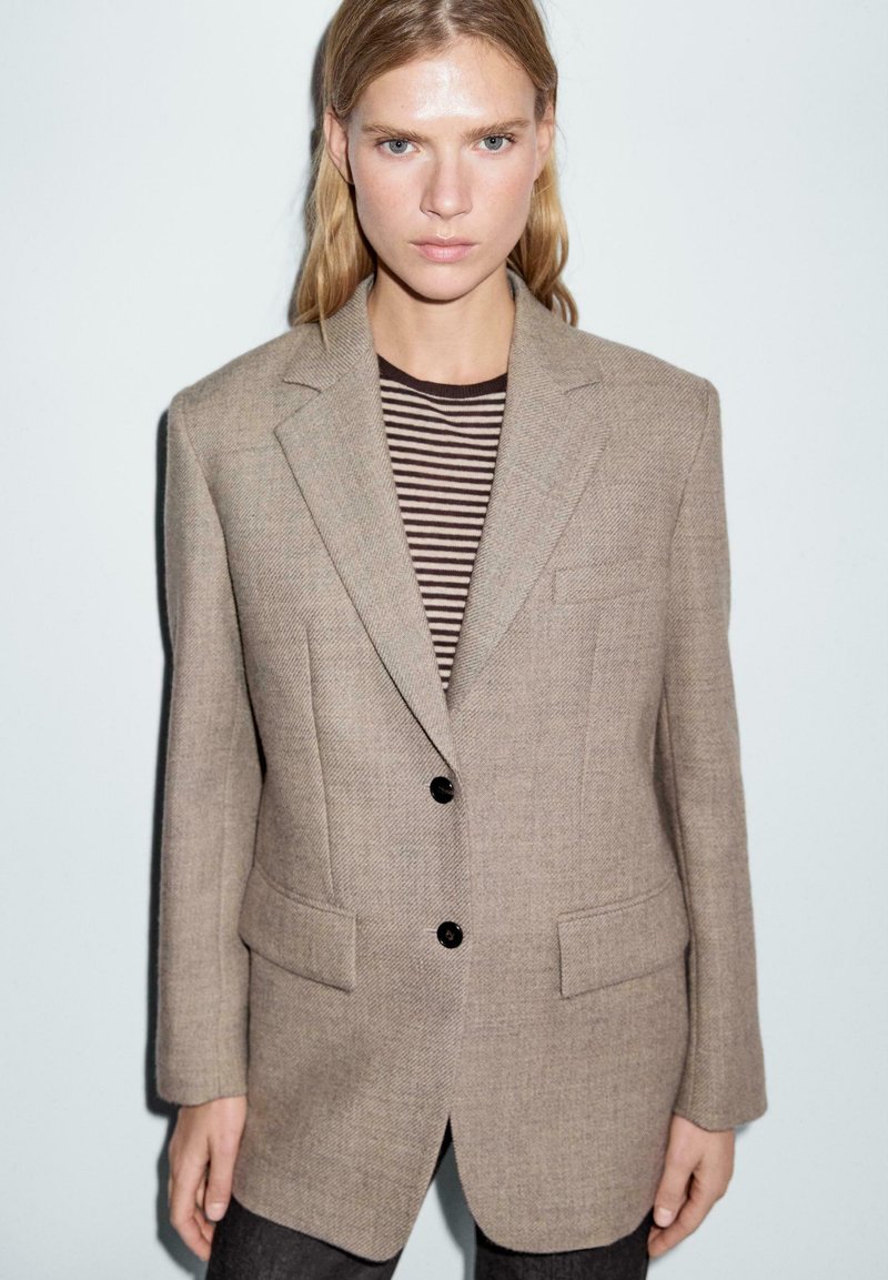 Blazer in lana beige con spalle strutturate, rever a punta e due tasche frontali. Indossato sopra una camicia a righe scure, con bottoni neri.