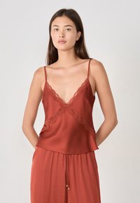 Modelo vistiendo una blusa sin mangas de color óxido con detalles de encaje, combinada con pantalones a juego, de pie contra un fondo sencillo.