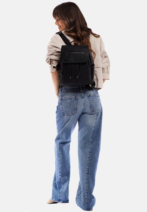 Schwarzer Rucksack aus strukturiertem Leder, mit Klappenverschluss, vorderer Reißverschluss-Tasche und verstellbaren Trägern, kombiniert mit hochgeschnittenen blauen Jeans.