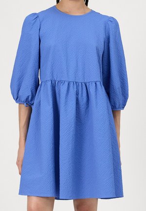 Frau trägt ein texturiertes, knielanges blaues Kleid mit aufgepufften Dreiviertelärmeln und geraffter Taille.