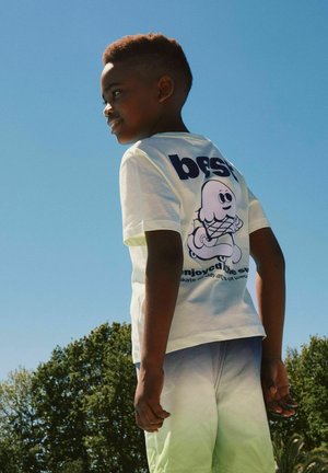 Boy with short hair wearing a white t-shirt with cartoon ice cream design and light green shorts, standing outdoors under clear sky.