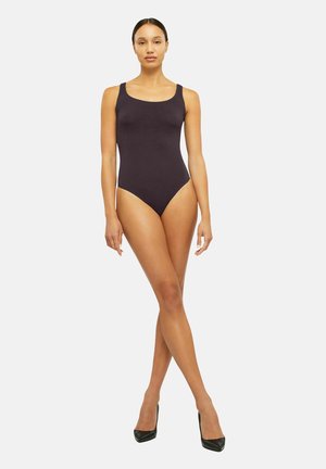Donna in piedi con le gambe incrociate che indossa un body nero senza maniche e tacchi neri su sfondo bianco.