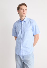 Camisa de manga corta azul claro de algodón, con frente de botones, un bolsillo de parche en el lado izquierdo del pecho y una textura sutil.