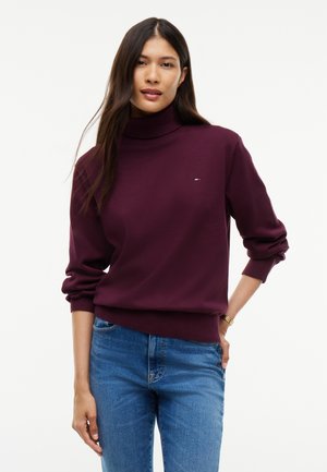 Kvinde med langt mørkt hår iført en burgundy rullekravetrøje og blå jeans, stående med den ene hånd på hoften.