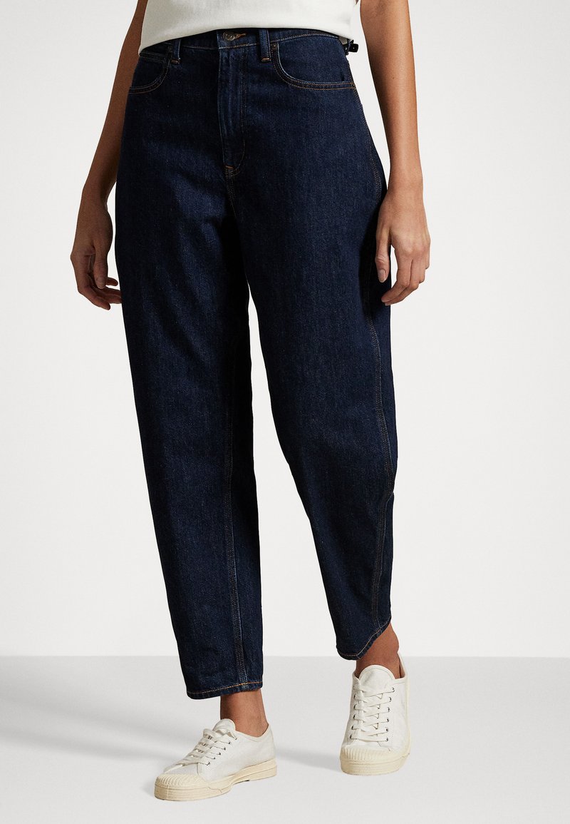 Mörkblå denimjeans med hög midja, smalnande ben, vit sömnad och avslappnade vita sneakers. Tyget ser robust ut med en slät yta.