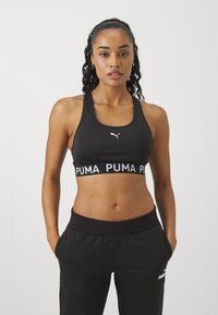 Crna sportska majica s širokim bijelim remenom "PUMA", s malim bijelim logotipom sprijeda. U kombinaciji s crnim trenerkama, prikazujući sportski izgled.