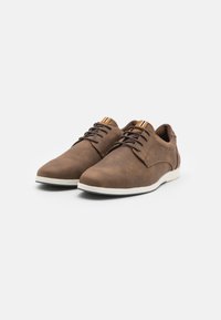 Zapatos marrones con cordones, de textura suave, suela blanca, punta redondeada y un acento a rayas en el talón. Fabricados en material sintético.