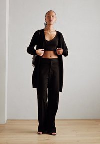 Un cardigan noir en tricot porté sur un top court noir et un pantalon taille haute, avec des sandales plates. Texture lisse, design minimaliste et coupe ajustée.
