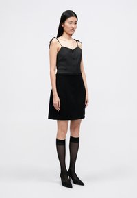 Μαύρη αμάνικη μπλούζα με κουμπιά και ιμάντες στους ώμους, συνδυασμένη με μια μαύρη φούστα. Το μοντέλο φοράει διάφανες μαύρες κάλτσες μέχρι το γόνατο και μαύρες γόβες με αιχμηρή μύτη.