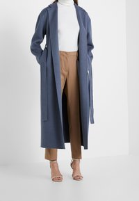 Manteau en mélange de laine bleu avec un devant ouvert, des manches longues et des liens à la taille, associé à un pantalon beige ajusté et des sandales à talons nude.