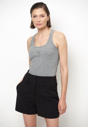 Vrouw met kort bruin haar, grijze mouwloze top en hoge zwarte shorts, staand tegen een witte achtergrond.