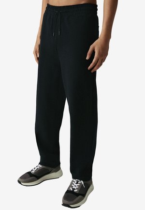 Man draagt zwarte joggingbroek met trekkoord en grijs-zwarte sneakers, staand tegen een effen lichte achtergrond.