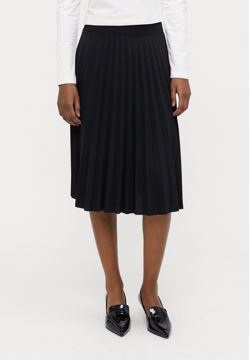 Femme portant une jupe midi plissée noire, un haut blanc à manches longues et des chaussures noires à bout pointu brillant, debout sur un fond blanc uni.