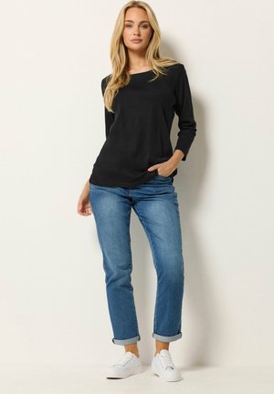 Eine Frau steht vor einer weißen Wand und trägt ein schwarzes Langarmshirt, hochgekrempelte blaue Jeans und weiße Turnschuhe, mit einer Hand in der Tasche.