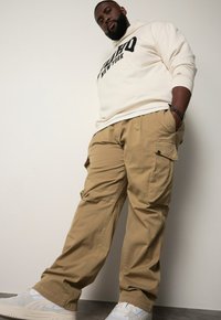 Beige cargobroeken met meerdere zakken, gecombineerd met een witte hoodie met zwarte letters. De outfit wordt compleet gemaakt met witte sneakers.