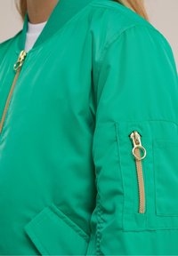 Grön bomberjacka med en slät textur, ficka med dragkedja på ärmen och guldtonade detaljer. Har ribbade krage och ärmslut.