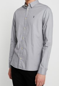 Mężczyzna w jasnoszarej koszuli z długim rękawem, zapinanej na guziki z kołnierzykiem, oraz czarnych spodniach, stojący na jednolitym tle.