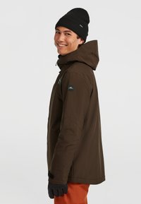Bordeaux geïsoleerde jas met capuchon, zwarte gebreide beanie en zwarte handschoenen. Bevat een logopatch op de schouder en oranje broek.
