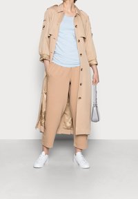Béžový trenčkot s knoflíky, světle modrý top, béžové cropped kalhoty a bílé tenisky, držící malou stříbrnou tašku.