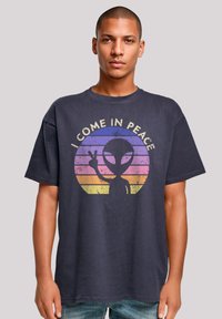 Navy blue cotton t-shirt featuring a graphic design of an alien with the text "I COME IN PEACE" and a sunburst in pink and yellow hues.