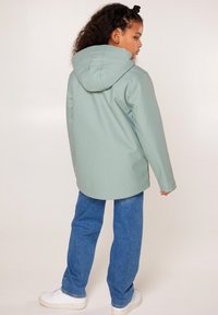 Giacca impermeabile verde chiaro con cappuccio, caratterizzata da una vestibilità comoda e una texture liscia, indossata sopra jeans blu a gamba dritta e sneakers bianche.