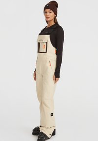 Beige sneeuwoverall met zwarte afwerking, verstelbare banden, meerdere zakken en oranje ritsaccenten. Zwarte skibotten maken de outfit compleet.