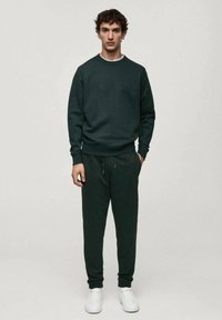 Temno zelena majica s kapuco z rebrastimi manšetami in ovratnikom, v kombinaciji z usklajenimi hlačami za prostočasne aktivnosti, ki imajo elastičen pas in vrvico.