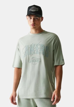 Joven con una camiseta oversized de color verde claro de Los Angeles Lakers y gorra negra de los Lakers, de pie frente a un fondo blanco.