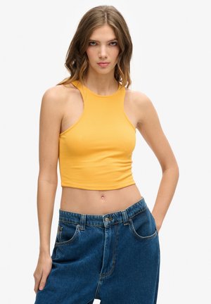 Jeune femme aux cheveux châtain clair portant un crop top jaune sans manches et un jean bleu ample, debout devant un fond uni.