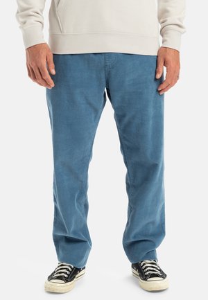 Mand iført lyseblå kordbuksere, sort-hvide sneakers og en lysebeige sweatshirt, stående mod en hvid baggrund.