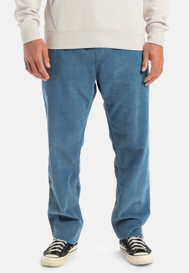 Homme portant un pantalon en velours côtelé bleu clair, des baskets noires et blanches, et un sweat-shirt beige clair, debout sur un fond blanc.
