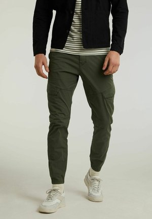 Man in olijfgroene cargobroek, witte sneakers, beige sokken, gestreept shirt en zwarte jas staand tegen een effen achtergrond.
