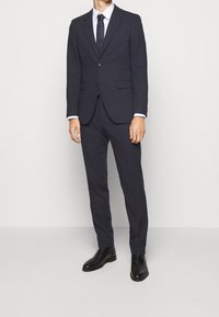 Marineblauer Anzug mit maßgeschneiderter Passform, bestehend aus einem Zwei-Knopf-Jacket, Reverskragen und Hosen mit flachem Front, kombiniert mit einem weißen Hemd und einer dunklen Krawatte. Schwarze Lederstiefel.