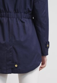 Veste bleu marine avec une ceinture élastique, des manches longues ornées de boutons dorés, et une texture lisse et mate.
