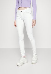 Mujer vestida con pantalones ajustados blancos de cintura alta, zapatillas blancas de suela gruesa y un suéter corto de botones lavanda, de pie contra un fondo sencillo.