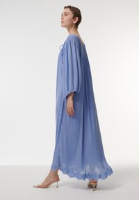 Vestido maxi plisado de color azul claro con mangas abullonadas, escote fruncido y borde de encaje en el dobladillo. Se lleva con sandalias de tacón transparentes.