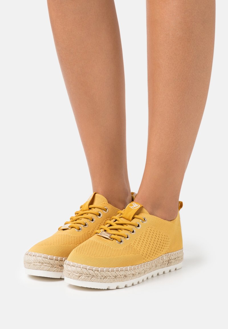 Chaussures décontractées jaunes à lacets avec semelles plateforme tissées style espadrille portées pieds nus sur fond blanc.