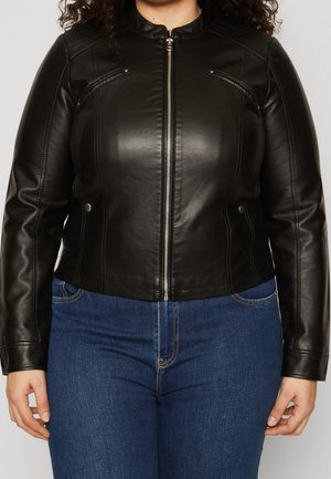 Frau trägt eine taillierte schwarze Lederjacke mit Reißverschluss vorne und blaue Jeans, steht vor einem schlichten hellen Hintergrund.