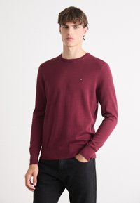 Burgunderfarget genser med rund hals laget av lett stoff med lange ermer. Har en liten logo på brystet og ribbestrikkede mansjetter og nederkant.