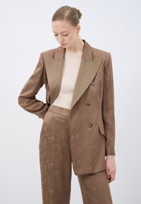 Blazer marrone con texture, colletto in contrasto di un marrone chiaro, chiusura doppiopetto con bottoni e pantaloni abbinati con motivo.