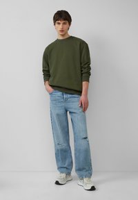 Olive green sweatshirt, luźny krój, prążkowane mankiety; jasnoniebieskie dżinsy z przetarciami; białe sneakersy z akcentami. Prosty design, casualowy wygląd.