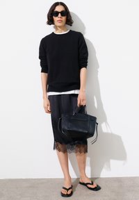Personne portant un pull noir par-dessus une chemise blanche, une jupe noire bordée de dentelle, des sandales noires, des lunettes de soleil, tenant un sac à main noir contre un mur blanc.