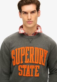 Szary sweatshirt z pomarańczowym haftowanym logo "SUPERDRY STATE". Nałożony na czerwoną i białą koszulę w kratę. Prosty, casualowy design.