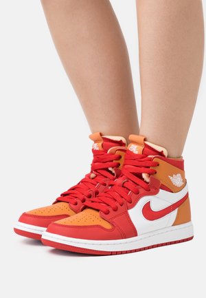 Jambes portant des baskets montantes Nike Air Jordan rouges, orange et blanches avec des lacets rouges sur un fond clair.