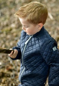Chaqueta acolchada azul marino con cierre de cremallera y cuello acanalado. Niño sosteniendo un palo, con cabello texturizado y expresión neutral. Fondo natural.