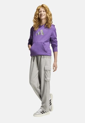 Joven con cabello rizado y rojo que lleva una sudadera con capucha morada de Adidas, pantalones cargo grises y zapatillas blancas, de pie relajado.